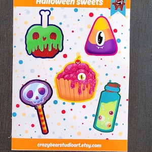 May include: A sticker sheet with Halloween-themed illustrations of sweets. Designs include a poison apple, a candy corn with one eye, a cupcake with an eyeball, a skull lollipop, and a potion bottle with eyeballs. The text "Halloween sweets" is at the top.