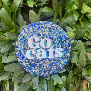 Puede incluir: Un posavasos redondo, azul y plateado con purpurina, con las palabras "Go Cats" en letras blancas.