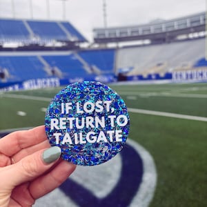 Puede incluir: Una pegatina redonda azul y blanca cubierta de purpurina con el texto "Si se pierde, vuelva al tailgate".