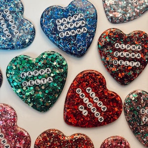 Puede incluir: Una colección de piezas de resina con forma de corazón y purpurina en varios colores, incluyendo azul, verde, rojo y rosa. Cada corazón presenta cuentas de letras blancas que deletrean letras griegas, posiblemente para hermandades.