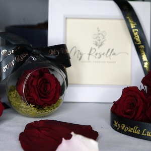 Può includere: Una singola rosa rossa conservata in una cupola di vetro con un nastro nero legato intorno. Il nastro ha la scritta dorata "My Rosella Luxury Forever Rose".