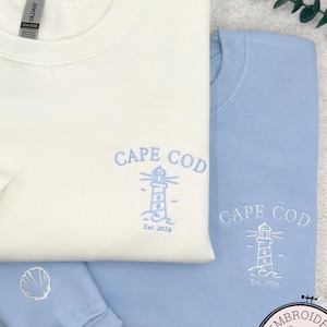 May include: Two sweatshirts, one cream and one light blue, are displayed. Both have the words "CAPE COD" and a lighthouse design embroidered in light blue. A seashell is embroidered on the cream sweatshirt's sleeve. The sweatshirts are folded on a white, textured surface.