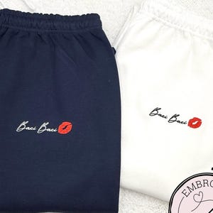 May include: Two pairs of sweatpants, one navy blue and one white, are displayed. Each pair features the embroidered text "Baci Baci" with a red lip graphic. The sweatpants have a drawstring waistband.