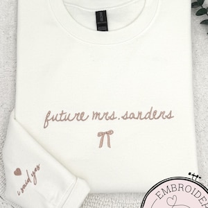 May include: Cream-colored sweatshirt with the embroidered text "future mrs. sanders" and a bow. The sleeve features "i said yes" and a heart. A circular logo with "Embroidery Design" is in the lower right corner.
