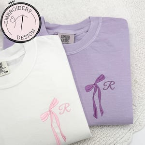 May include: Two t-shirts with embroidered bows and the letter "R" on them. The t-shirt on the left is white with a pink bow and the t-shirt on the right is lavender with a purple bow. The t-shirts are on a white crocheted surface.