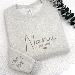May include: A gray sweatshirt with the word "Nana" embroidered in brown cursive lettering. The text "EST. 2025" is embroidered below the word "Nana" with a small heart symbol. The words "Eliza Abigail" are embroidered on the left sleeve in brown cursive lettering.