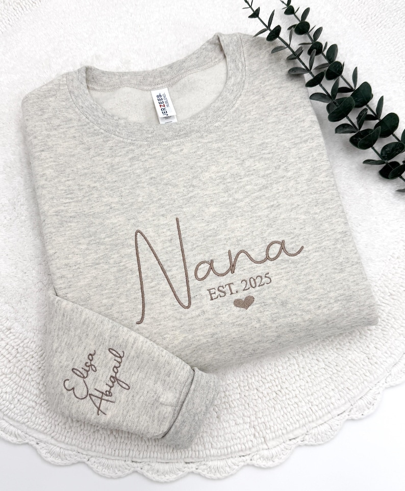 May include: A gray sweatshirt with the word "Nana" embroidered in brown cursive lettering. The text "EST. 2025" is embroidered below the word "Nana" with a small heart symbol. The words "Eliza Abigail" are embroidered on the left sleeve in brown cursive lettering.