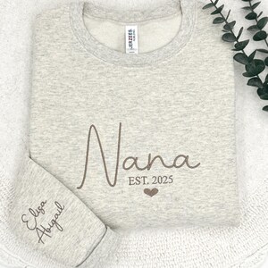 May include: A gray sweatshirt with the word "Nana" embroidered in brown cursive lettering. The text "EST. 2025" is also embroidered below the word "Nana" with a small heart symbol. The sweatshirt has a small tag on the left shoulder that says "JERZEES" and "MADE IN MEXICO". The sweatshirt is folded in half and the bottom of the sweatshirt is visible. The text "Eliza Abigail" is embroidered in brown cursive lettering on the bottom of the sweatshirt.