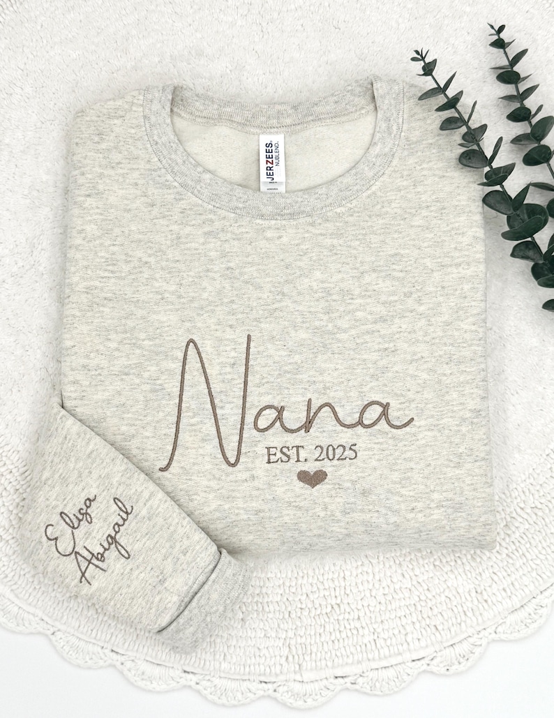 May include: A gray sweatshirt with the word "Nana" embroidered in brown cursive lettering. The text "EST. 2025" is also embroidered below the word "Nana" with a small heart symbol. The sweatshirt has a small tag on the left shoulder that says "JERZEES" and "MADE IN MEXICO". The sweatshirt is folded in half and the bottom of the sweatshirt is visible. The text "Eliza Abigail" is embroidered in brown cursive lettering on the bottom of the sweatshirt.