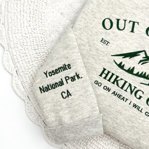 May include: A light gray sweatshirt with green text that reads "OUT", "HIKING", and "Yosemite National Park, CA". The sweatshirt has a ribbed cuff and a mountain graphic.