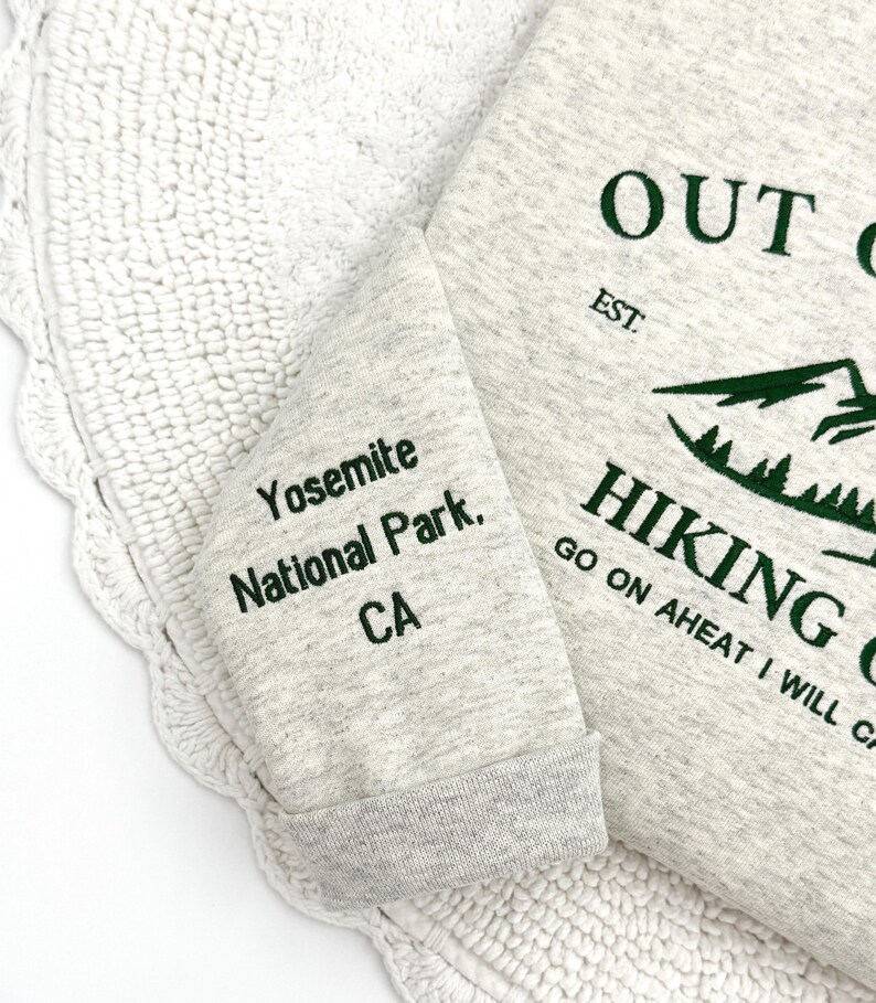 May include: A light gray sweatshirt with green text that reads "OUT", "HIKING", and "Yosemite National Park, CA". The sweatshirt has a ribbed cuff and a mountain graphic.