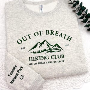 May include: A light gray sweatshirt with the text "OUT OF BREATH HIKING CLUB" in dark green. The design includes a mountain graphic and the text "Yosemite National Park, CA" on the sleeve. The sweatshirt is folded.