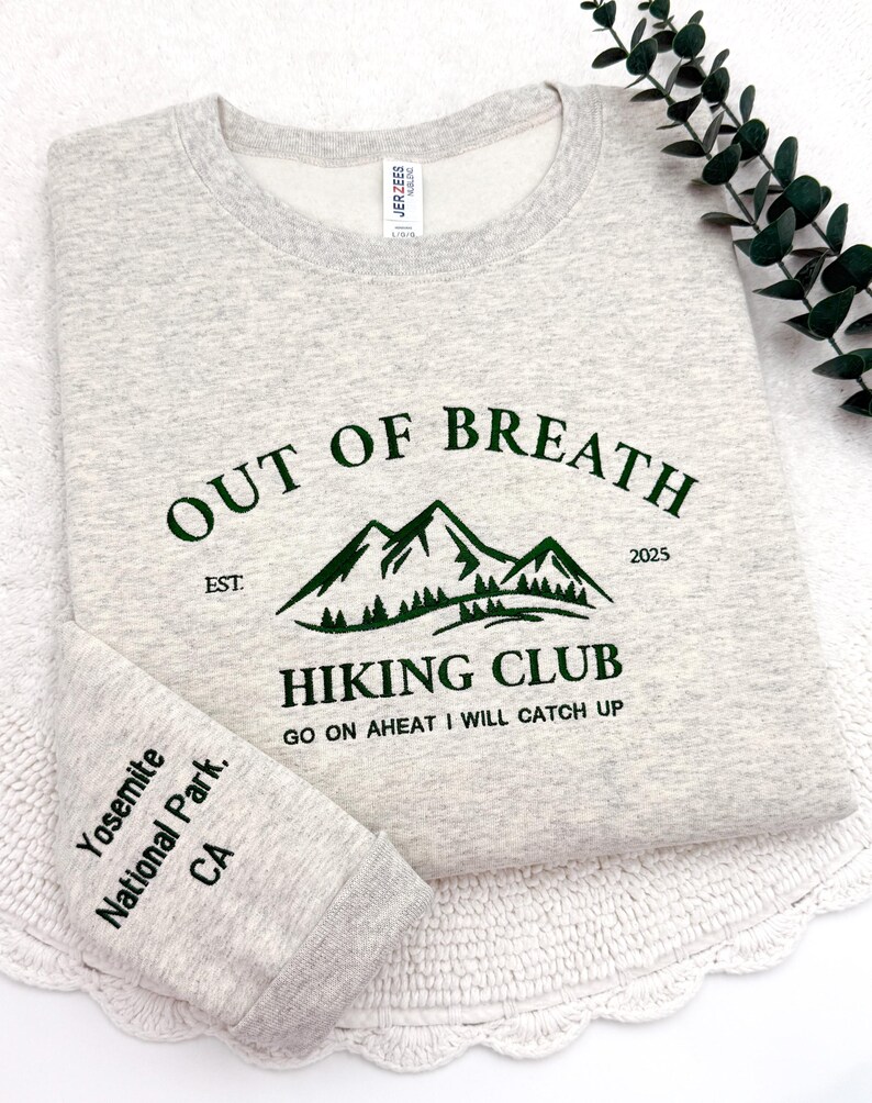 May include: A light gray sweatshirt with the text "OUT OF BREATH HIKING CLUB" in dark green. The design includes a mountain graphic and the text "Yosemite National Park, CA" on the sleeve. The sweatshirt is folded.