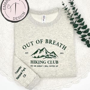 May include: A light gray sweatshirt with a green mountain graphic and the text "OUT OF BREATH HIKING CLUB". The sleeve reads "Yosemite National Park, CA". The sweatshirt has a crew neck and is made of a soft material.