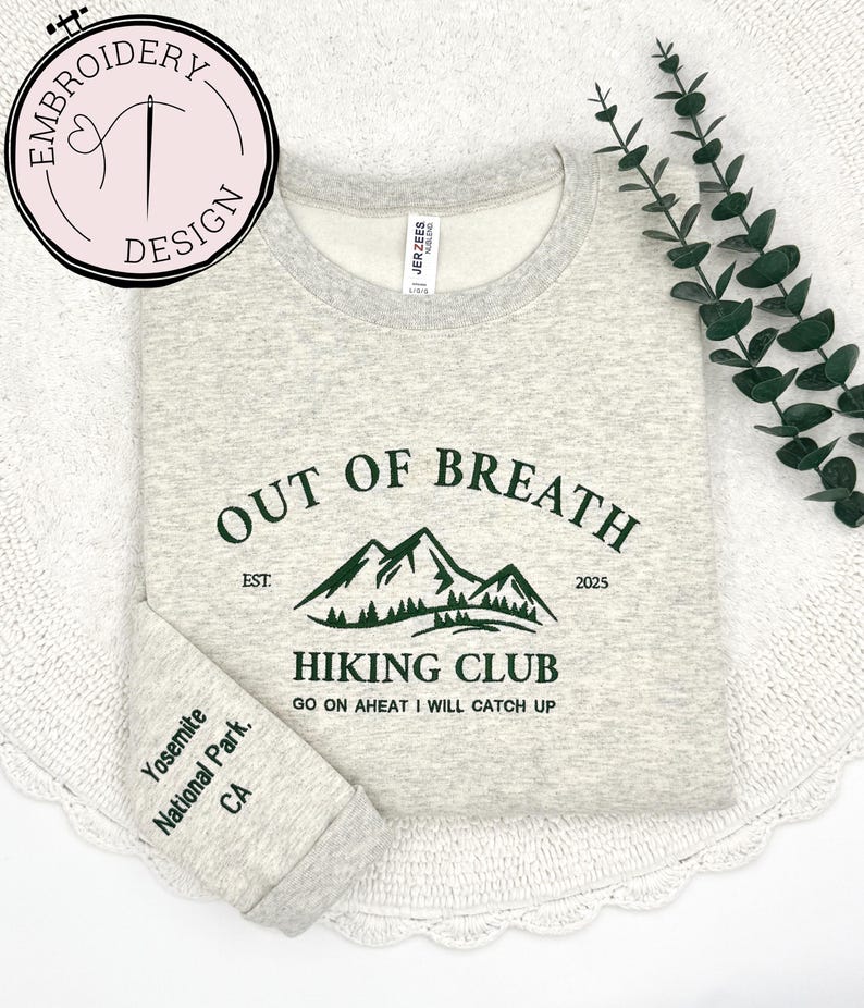 May include: A light gray sweatshirt with a green mountain graphic and the text "OUT OF BREATH HIKING CLUB". The sleeve reads "Yosemite National Park, CA". The sweatshirt has a crew neck and is made of a soft material.
