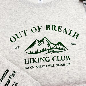 May include: A light gray sweatshirt with the text "OUT OF BREATH HIKING CLUB" in dark green. The design includes mountain graphics and the text "GO ON AHEAD I WILL CATCH UP". The sleeve reads "Yosemite National Park, CA".