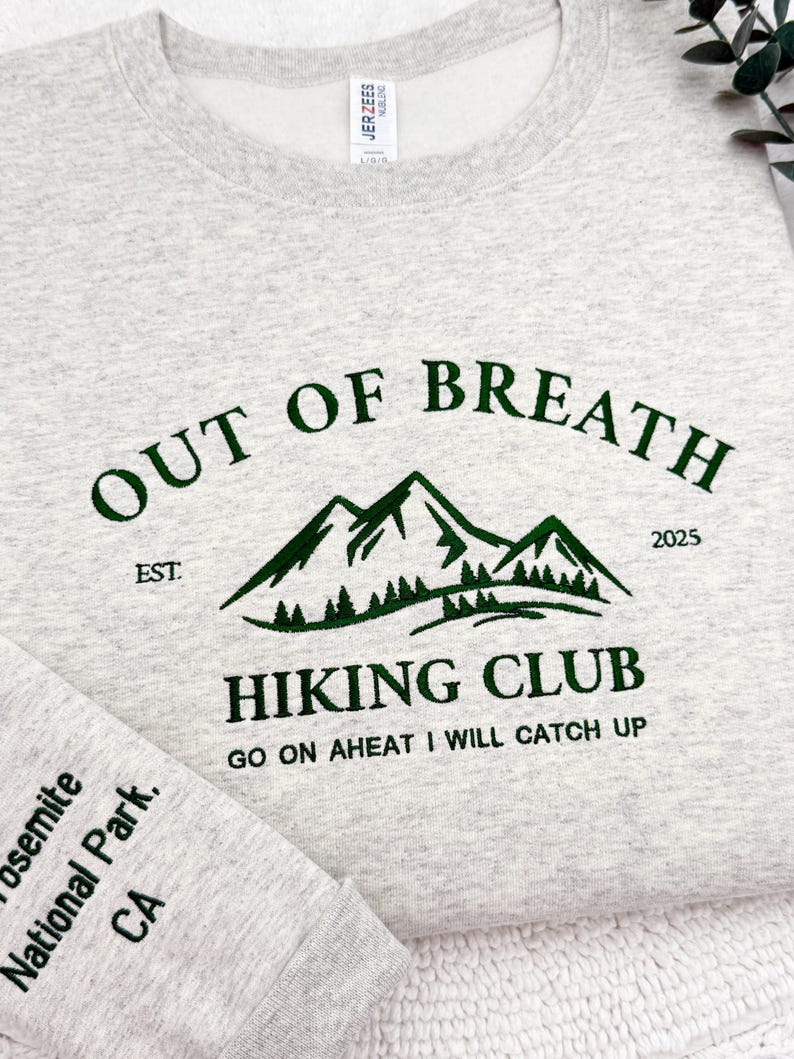 May include: A light gray sweatshirt with the text "OUT OF BREATH HIKING CLUB" in dark green. The design includes mountain graphics and the text "GO ON AHEAD I WILL CATCH UP". The sleeve reads "Yosemite National Park, CA".