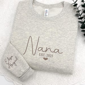 May include: Gray sweatshirt with the word "Nana" embroidered in brown script with the text "EST. 2025" below. The left sleeve has the names "Elisa" and "Abigail" embroidered in brown script.