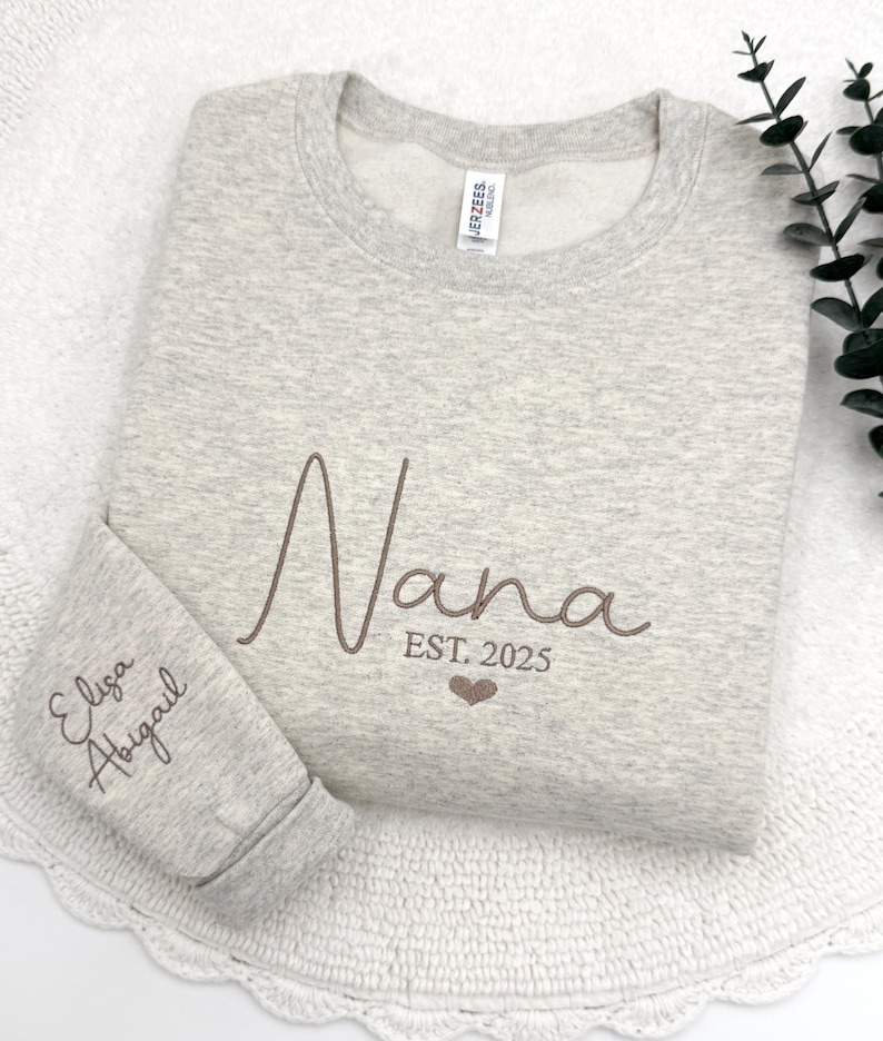 May include: Gray sweatshirt with the word "Nana" embroidered in brown script with the text "EST. 2025" below. The left sleeve has the names "Elisa" and "Abigail" embroidered in brown script.