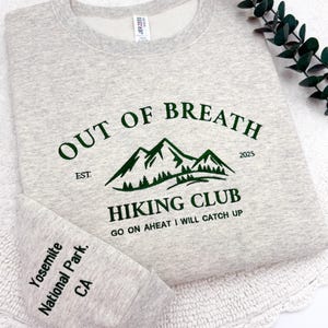 May include: A light gray sweatshirt with the text "OUT OF BREATH HIKING CLUB" in green, featuring a mountain graphic. The sleeve cuff reads "Yosemite National Park, CA". The sweatshirt has a crew neck.