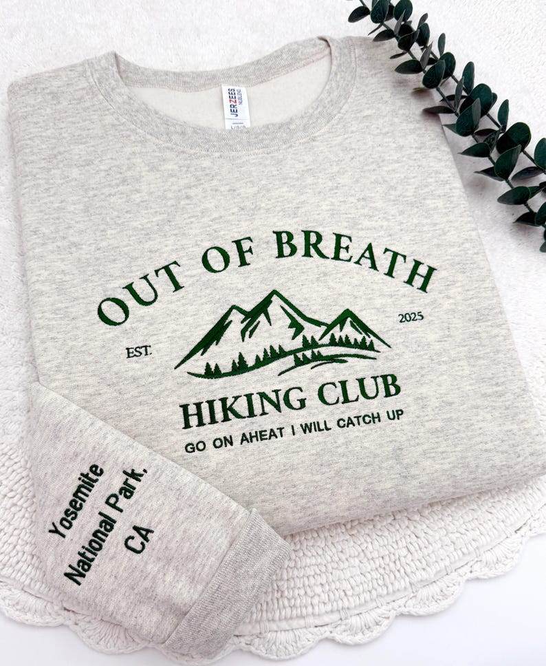 May include: A light gray sweatshirt with the text "OUT OF BREATH HIKING CLUB" in green, featuring a mountain graphic. The sleeve cuff reads "Yosemite National Park, CA". The sweatshirt has a crew neck.