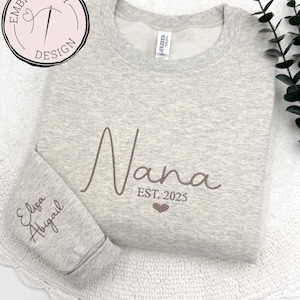 May include: A gray sweatshirt with the word "Nana" embroidered in brown on the front. The text "EST. 2025" is embroidered below the word "Nana". The names "Elisa" and "Abigail" are embroidered on the left sleeve.