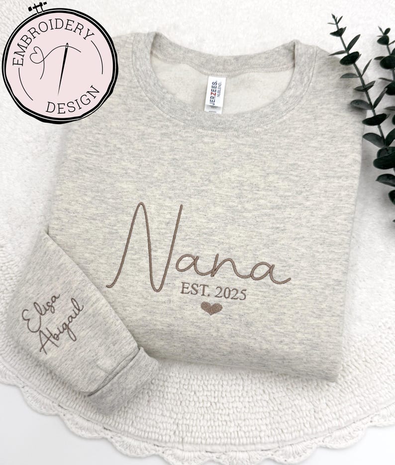May include: A gray sweatshirt with the word "Nana" embroidered in brown on the front. The text "EST. 2025" is embroidered below the word "Nana". The names "Elisa" and "Abigail" are embroidered on the left sleeve.