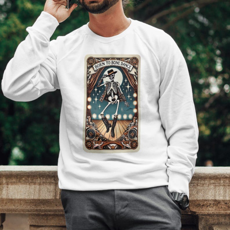 May include: White long-sleeve sweatshirt with a graphic of a skeleton dancing on a stage, wearing a hat and boots. The graphic has the text "Born to Bone Dance" at the top. The design features a vintage, theatrical style.