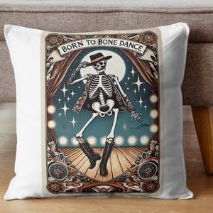 May include: White square throw pillow featuring a skeleton in a hat and jacket dancing on a stage. The pillow has the text "BORN TO BONE DANCE" across the top. The design includes a full moon, stars, and stage curtains.