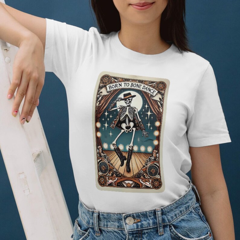 May include: White t-shirt featuring a dancing skeleton graphic with the text "Born to Bone Dance." The design includes a skeleton in a hat and coat, set against a stage backdrop with a full moon and stars. The graphic is framed by an ornate border.