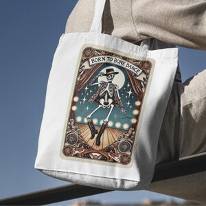 May include: White tote bag featuring a vintage-style illustration of a dancing skeleton wearing a hat and boots. The image includes the text "BORN TO BONE DANCE" above the skeleton. The background has a stage setting with a full moon and stars.