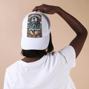 May include: White trucker hat with a mesh back and a front panel featuring a dancing skeleton illustration. The artwork includes the text "Born to Bone Dance" in a banner above the skeleton. The hat is a classic accessory.