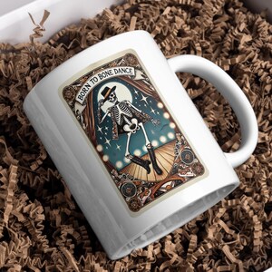 May include: White ceramic mug featuring a dancing skeleton illustration with the text "Born to Bone Dance." The design includes a skeleton in a hat and boots, set against a backdrop of a full moon and stars, framed by ornate borders.