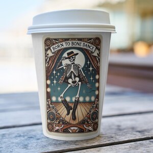 May include: White coffee cup with a lid, featuring a decorative sticker. The sticker shows a skeleton in a hat and boots, dancing on a stage with the text "Born to Bone Dance" above. The design includes a full moon and stage curtains.