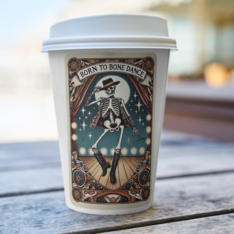 May include: White coffee cup with a lid, featuring a decorative sticker. The sticker shows a skeleton in a hat and boots, dancing on a stage with the text "Born to Bone Dance" above. The design includes a full moon and stage curtains.