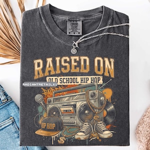 May include: Dark gray Comfort Colors t-shirt with a graphic design. The design features the text "RAISED ON OLD SCHOOL HIP HOP" above a boombox, microphone, cap, sneakers, and a vinyl record. A silver necklace is draped across the shirt.