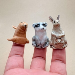 May include: Three small ceramic animal figurines designed as finger puppets. The left figure is a tan seal, the center figure is a grey and white patterned bear, and the right figure is a brown kangaroo with a small joey in its pouch.