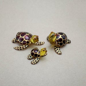 Puede incluir: Tres figuras de tortugas de cristal. Las tortugas tienen un patrón de manchas marrones y doradas en sus caparazones y aletas. Las cabezas son amarillas con ojos rojos. Las tortugas son de diferentes tamaños.