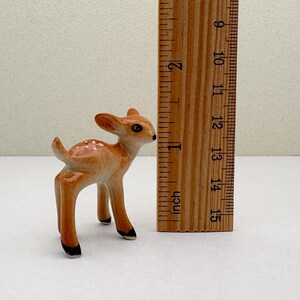 May include: A small ceramic fawn figurine, approximately 2 inches tall, with a light brown coat and white spots. The figurine has black hooves and a detailed, alert expression. A wooden ruler is next to the fawn.
