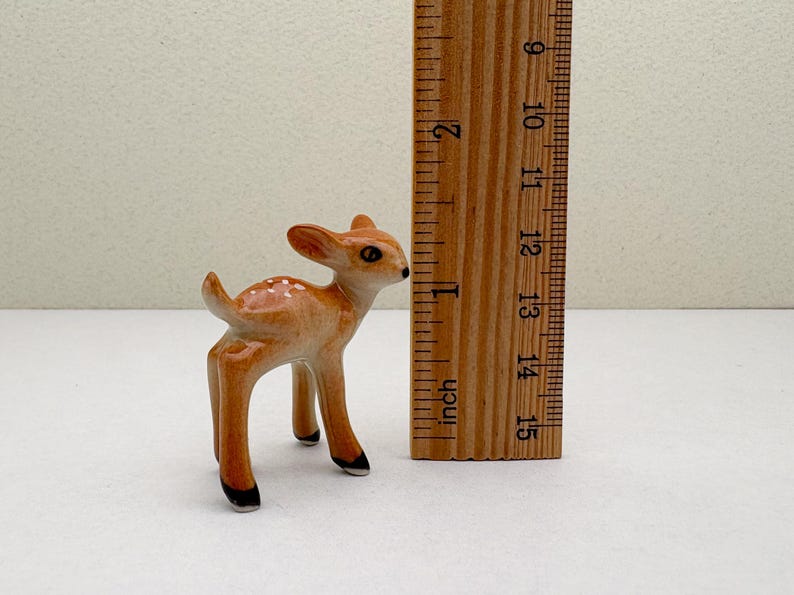 May include: A small ceramic fawn figurine, approximately 2 inches tall, with a light brown coat and white spots. The figurine has black hooves and a detailed, alert expression. A wooden ruler is next to the fawn.