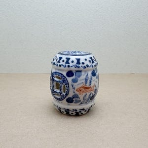 Puede incluir: Pequeño taburete cilíndrico de cerámica con base blanca y detalles azules. El diseño incluye un pez, plantas acuáticas y un recorte en forma de moneda. El taburete mide unos 20 cm de alto.