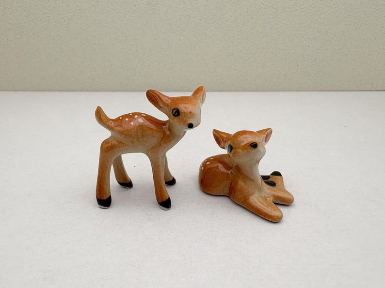 May include: Two ceramic deer figurines. One deer is standing, the other is lying down. Both are light brown with white spots and black hooves. The figurines have a glossy finish and are set against a plain background.