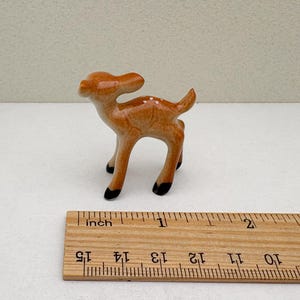 May include: A small ceramic deer figurine, standing on four legs with black hooves. The deer is light brown with white spots and a small tail. A wooden ruler with inch markings is in the foreground.