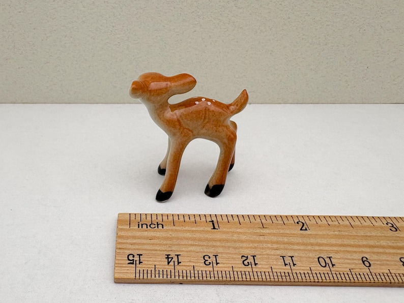 May include: A small ceramic deer figurine, standing on four legs with black hooves. The deer is light brown with white spots and a small tail. A wooden ruler with inch markings is in the foreground.