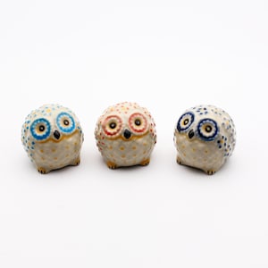 Könnte beinhalten: Drei Keramik-Eulenfiguren mit weißen Körpern und bunten Akzenten. Die Eulen haben jeweils blaue, rote und blaue Augen. Die Eulen sind alle nach vorne gerichtet und haben ein gepunktetes Muster auf ihren Körpern.