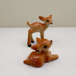 May include: Two ceramic deer figurines. One deer is standing, the other is lying down. Both are light brown with white spots and black accents on the hooves and eyes. The figurines are on a white surface.