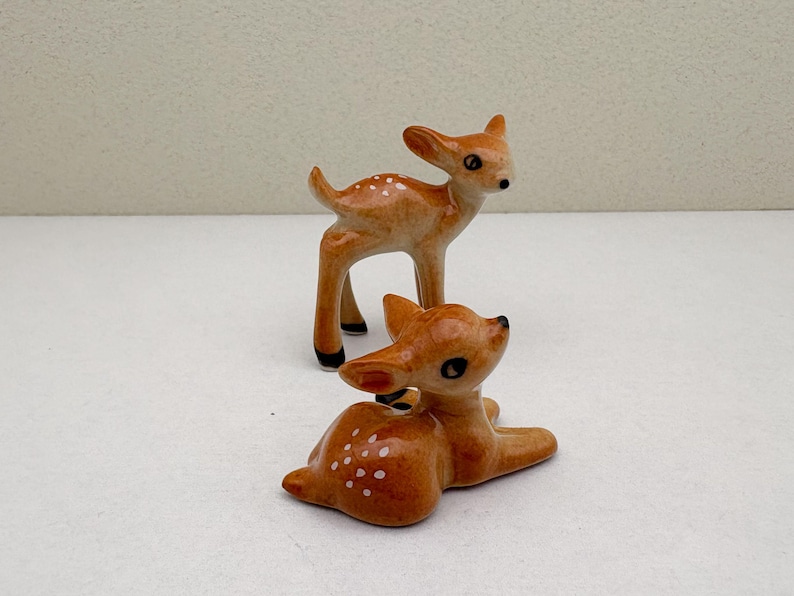 May include: Two ceramic deer figurines. One deer is standing, the other is lying down. Both are light brown with white spots and black accents on the hooves and eyes. The figurines are on a white surface.