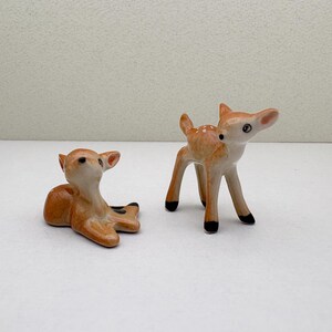 May include: Two ceramic deer figurines. One is lying down, the other is standing. Both are light brown with white spots and black hooves. The deer have detailed features, including ears and eyes, and are set against a white background.