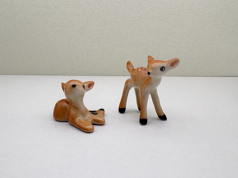 May include: Two ceramic deer figurines. One is lying down, the other is standing. Both are light brown with white spots and black hooves. The deer have detailed features, including ears and eyes, and are set against a white background.