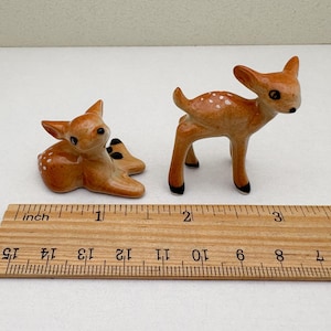 May include: Two ceramic deer figurines. One is lying down, the other is standing. Both are brown with white spots and black accents. A wooden ruler with inch markings is in the foreground.